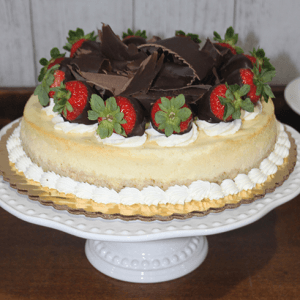 The GD-001 New York Style Cheesecake, topped with chocolate shavings and chocolate-dipped strawberries and decorated with whipped cream, is displayed on a scalloped-edge white cake stand.
