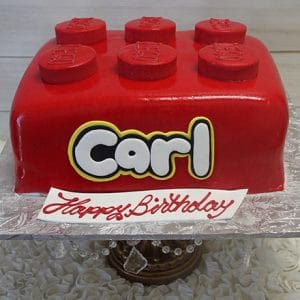 The CB-004D is a red LEGO brick-shaped cake with six round bumps, Carl’s name in bold letters, and a "Happy Birthday" sign in red icing. The cake is displayed on a decorated stand.