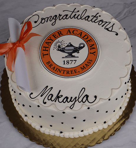 A round white cake with scalloped icing, black dots, a diploma and orange ribbon, reading "Congratulations Makayla" with a Thayer Academy 1877 Braintree, Mass emblem. Product: GC-001L Thayer Academy.