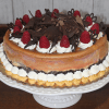 The GD-004 Raspberry Chambord Cheesecake is topped with whipped cream, fresh raspberries, chocolate shavings, and chocolate decorations, displayed on a white cake stand.