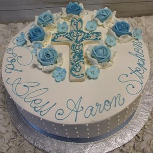 RC-053: Round white cake featuring blue and white flowers, a blue ornate cross topper, and "God Bless Aaron Stephen" in blue icing around the edge. Presented on a silver board.