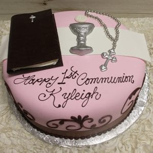 The RC-073 is a pink and white cake featuring a Bible, chalice, rosary, and “Happy 1st Communion Kyleigh” in brown icing. Presented on a silver cake board.