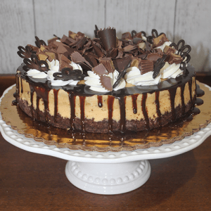 The GD-012 Peanut Butter Cheesecake sits on a white cake stand, topped with whipped cream, chocolate shavings and curls, and drizzled with chocolate sauce.