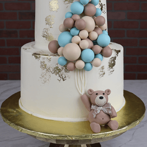 The TC-000J cake features two white tiers with gold accents, blue and beige balloon clusters on top, and a fondant teddy bear holding balloon strings at the base. Presented on a gold cake stand.