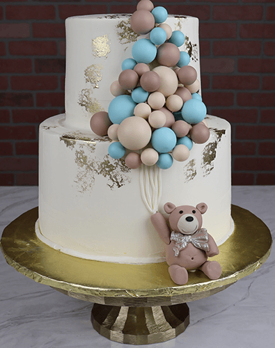 The TC-000J cake features two white tiers with gold accents, blue and beige balloon clusters on top, and a fondant teddy bear holding balloon strings at the base. Presented on a gold cake stand.