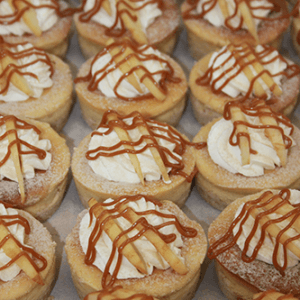 Rows of CO-021 3" Caramel Apple Cheesecakes topped with whipped cream, thin apple slices, and a caramel drizzle.