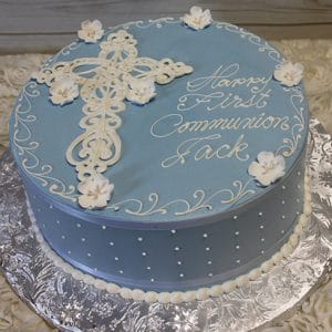 The RC-015 cake is round and blue with white dots, a white icing cross, small white flowers, and "Happy First Communion Jack" written in white icing. It is displayed on a silver board.