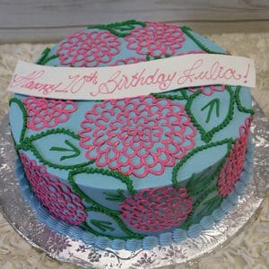 BD-147 is a round blue cake featuring pink and green floral designs, a white banner with "Happy 17th Birthday Julia!" in pink script, and is presented on a silver board.