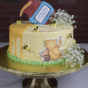 The BB-000D is a round cake with yellow icing, honey drip, buzzing bees, a fondant "Hunny" pot and flowers on top, plus Winnie the Pooh and Piglet artwork on the side. Served on a gold stand.