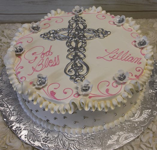 RC-022 is a round white cake with a silver cross, small white flowers, silver dots, ruffled edges, and “God Bless Lillian” in pink writing. It’s presented on an embossed silver board.