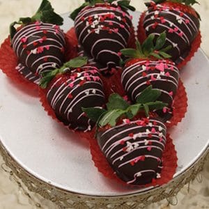 Six VC-031 Chocolate dipped strawberries with pink and white drizzle and heart-shaped sprinkles are arranged on a silver stand, each in a red paper cup.