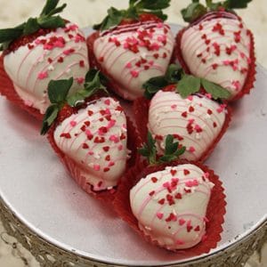VC-032 White chocolate dipped strawberries: Six strawberries coated in white chocolate, drizzled with pink icing, topped with red heart-shaped sprinkles, and presented in red paper cups on a silver platter.