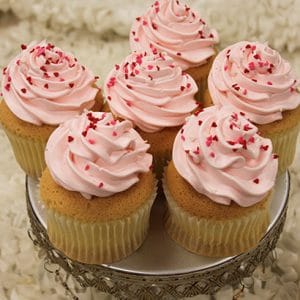 Six VC-038 Display Valentine gold cupcakes, filled with chocolate mousse and topped with light pink frosting and red heart-shaped sprinkles, are arranged on a decorative silver stand against a white textured background.