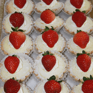 Rows of CO-077 New York Cheesecake mini tarts topped with whipped cream and whole strawberries, dusted with powdered sugar, and nestled in white paper wrappers are neatly arranged on a tray.