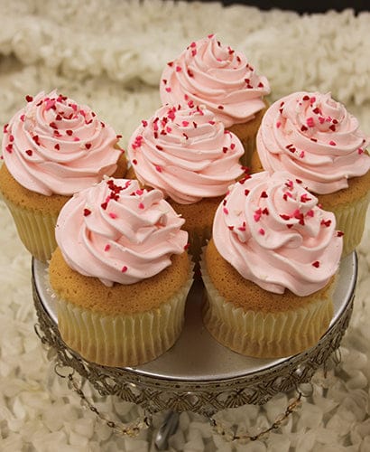 Six VC-038 Display Valentine gold cupcakes, filled with chocolate mousse and topped with light pink frosting and red heart-shaped sprinkles, are arranged on a decorative silver stand against a white textured background.