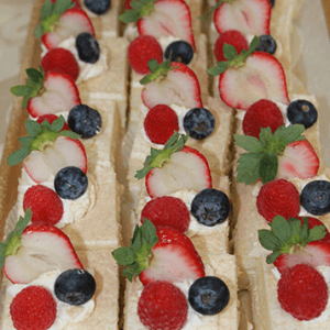 Rows of CO-072 Tres Leches Squares, topped with whipped cream, halved strawberries, blueberries, and raspberries, neatly arranged on a tray.