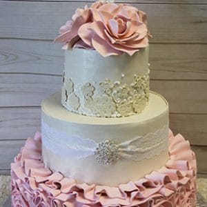 WD-015: A three-tiered white cake with pink ruffles, lace patterns, large fondant flowers, pearls, a brooch, and a white cross topper, displayed on a light wooden background.