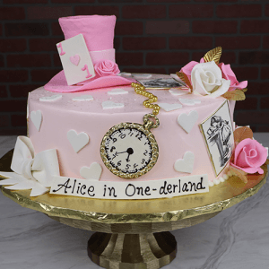The CB-039 Alice in Wonderland cake features a pink theme with a teacup, playing card, roses, gold leaves, white hearts, vintage images, and a gold pocket watch on a gold stand labeled “Alice in One-derland.”.