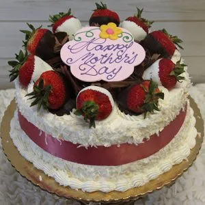 The MD-005 Pina Colada Cake is a round cake with white frosting, chocolate-covered strawberries on top, a pink ribbon around the side, and a pink plaque that says "Happy Mother’s Day" with a yellow flower decoration.