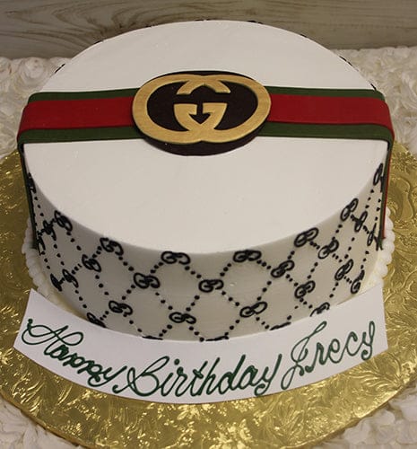 A round "BD-031 Designer Belt" cake with a gold logo, red and green stripe, black repeating patterns, and a front card saying “Happy Birthday Greg” in green script.