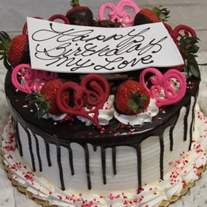 VC-009 is a round cake with white icing and chocolate drip, topped with strawberries, pink chocolate hearts, whipped cream, and a “Happy Birthday My Love” card. Pink sprinkles decorate the base.