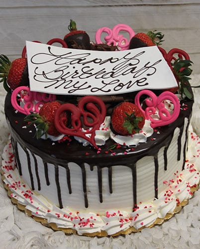 VC-009 is a round cake with white icing and chocolate drip, topped with strawberries, pink chocolate hearts, whipped cream, and a “Happy Birthday My Love” card. Pink sprinkles decorate the base.