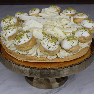 The GD-029B Gâteau St. Honoré with Pistachio is a round tart topped with cream puffs glazed in white, chopped pistachios, piped cream, and white chocolate shavings on a gold cake stand.