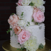 WD-001S is a two-tiered white cake with textured dots, topped with large, realistic pink, peach, and white flowers cascading down the side. It is presented on a gold cake board.