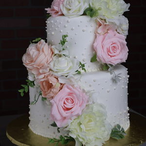 WD-001S is a two-tiered white cake with textured dots, topped with large, realistic pink, peach, and white flowers cascading down the side. It is presented on a gold cake board.