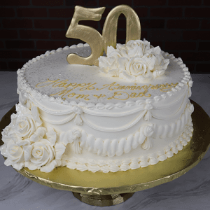 AN-023: White frosted cake with a gold “50” topper, white icing roses, gold script “Happy Anniversary Mom & Dad,” and gold board. Brick wall background.