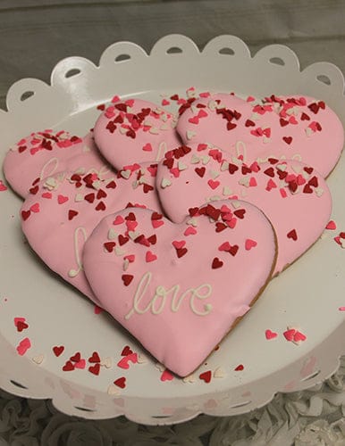 CE-020f Valentine's: Heart-shaped cookies with pink icing and "love," decorated with red, pink, and white heart sprinkles, arranged on a white scalloped plate.