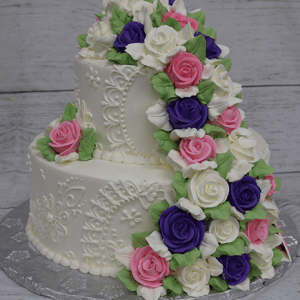 The TC-002B features a two-tiered white cake adorned with detailed piping and fondant roses in purple, pink, and white with green leaves, presented on a silver cake board against a rustic wood background.