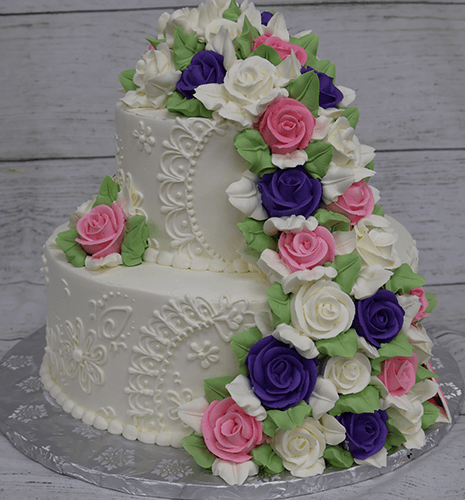The TC-002B features a two-tiered white cake adorned with detailed piping and fondant roses in purple, pink, and white with green leaves, presented on a silver cake board against a rustic wood background.
