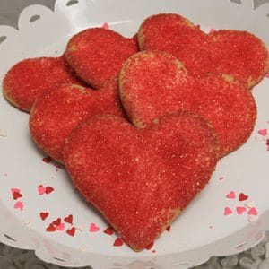 CE-020b Valentine's heart-shaped cookies with red sugar sprinkles are beautifully arranged on a white decorative plate, surrounded by small heart confetti.