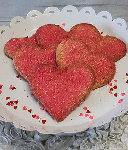 CE-020c Valentine's heart-shaped cookies with red-pink sugar sprinkles are arranged on a white scalloped tray, decorated with small heart confetti and set on a textured white surface.