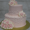 Two-tiered pink cake (TC-002C) featuring realistic pink and white sugar roses, white edible pearls, and set on a gold cake board against a rustic wood background.