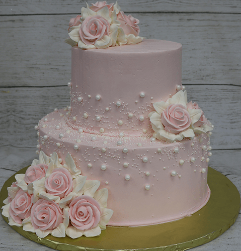 Two-tiered pink cake (TC-002C) featuring realistic pink and white sugar roses, white edible pearls, and set on a gold cake board against a rustic wood background.