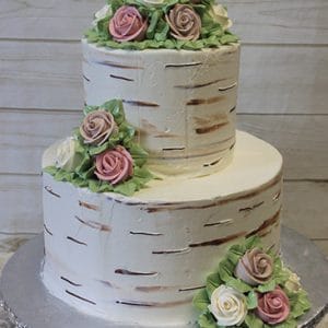 WC-004: Two-tiered birch bark-inspired cake, topped and accented with pink and white buttercream roses and green leaves, displayed on a silver cake board against a light wooden background.