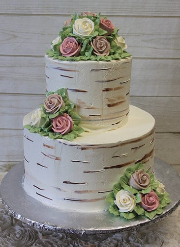 WC-004: Two-tiered birch bark-inspired cake, topped and accented with pink and white buttercream roses and green leaves, displayed on a silver cake board against a light wooden background.