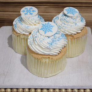 Three CH-061B Gold cupcakes with swirled white frosting, blue sprinkles, and white chocolate toppers featuring blue snowflakes are displayed on a white wooden board.
