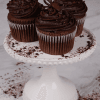 Three CC-000E Chocolate moca cupcakes with dark chocolate frosting and chocolate toppings sit on a white cake stand, surrounded by cocoa powder on a light marble surface.