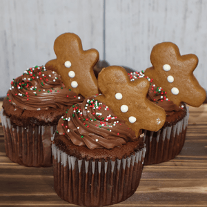 Three CH-062A Chocolate cupcakes with white filling are topped with chocolate frosting, multicolored sprinkles, and small gingerbread people cookies, shown on a wooden surface against a light wood background.