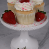 Three CC-000Q Strawberry Shortcake Cupcakes, topped with strawberries and pink sprinkles, sit on a white cake stand surrounded by fresh strawberries.