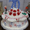 The BD-047 is a round birthday cake with red icing roses, a large blue 70 topper, golf-themed decor, red and blue writing, and a "Happy Birthday" message on white frosting.