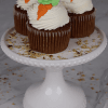 Three CC-000L carrot cupcakes with white frosting and carrot-shaped decorations are displayed on a white cake stand, with crumbs scattered around.