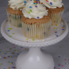 Three CC-000P Funfetti Cupcakes with white frosting and colorful sprinkles sit on a white cake stand, with extra sprinkles scattered around on a gray surface.