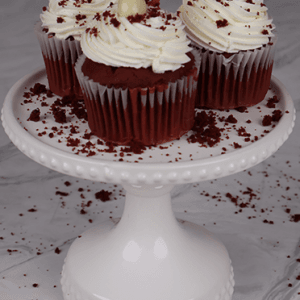 Three CC-000r red velvet cupcakes with white frosting, white chocolate balls, and red sprinkles sit on a white cake stand, red crumbs scattered around.