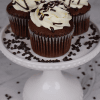 Three CC-006 Display Chocolate whipped cream Cupcakes with swirled white frosting and chocolate drizzle are on a white cake stand, surrounded by chocolate chips.