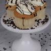 Three CC-007 Display Gold whipped cream Cupcakes with swirled white frosting and chocolate drizzle sit on a white cake stand, surrounded by scattered mini chocolate chips.