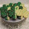 STP-001B St. Patrick's Day Sugar Cookies, shamrock-shaped and half dipped in chocolate with green sprinkles and lines, are arranged on a silver stand with bead accents on a white textured surface.